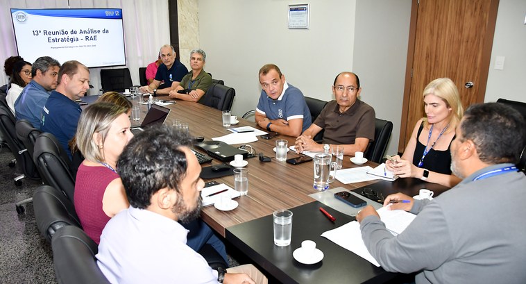 A imagem mostra uma reunião formal acontecendo em uma sala de conferências. Várias pessoas, home...