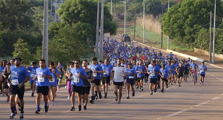 A imagem mostra uma grande corrida de rua realizada em uma avenida larga. Centenas de pessoas pa...