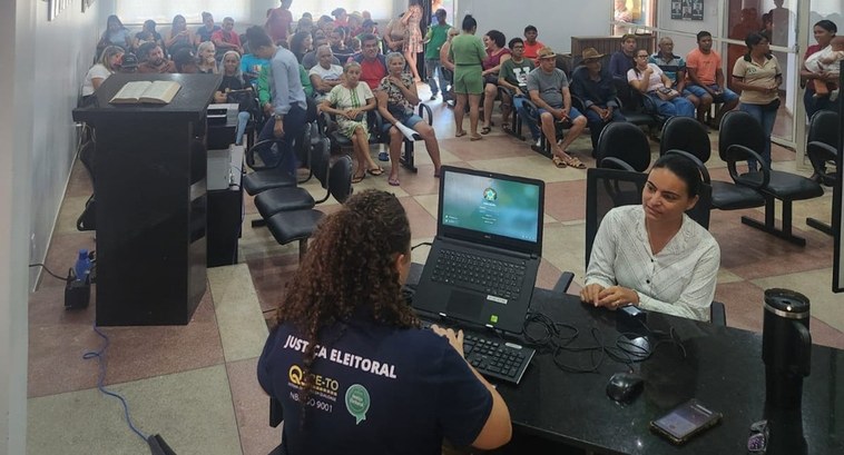 A imagem mostra o interior de um espaço público, com várias pessoas aguardando sentadas em cadei...