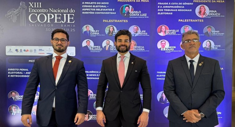 A imagem mostra os três juízes do TRE-TO em pé, posando para uma foto em frente a um painel inst...