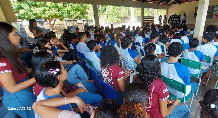 A imagem mostra uma grande audiência de jovens em um evento, em um ambiente escolar.