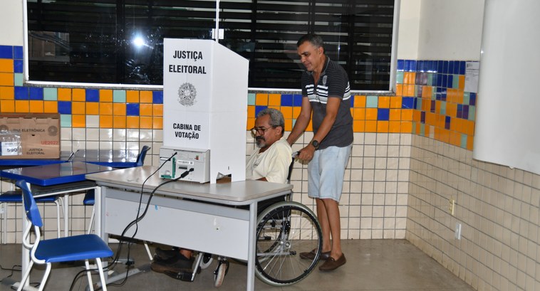 A imagem mostra um momento de votação em uma seção eleitoral. Um eleitor cadeirante está posicio...