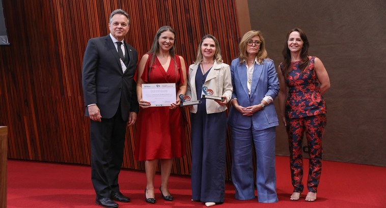 A imagem mostra cinco pessoas posando para uma foto em um ambiente formal. À esquerda, um homem ...