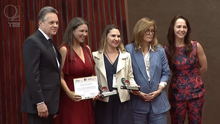 Justiça Eleitoral do Tocantins é premiada no TSE por ação que garantiu o voto de pessoas em situação de rua A imagem mostra cinco pessoas posando para uma foto em um ambiente formal. À esquerda, há um hom...