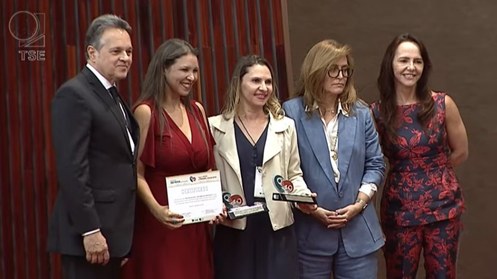 A imagem mostra cinco pessoas posando para uma foto em um ambiente formal. À esquerda, há um hom...