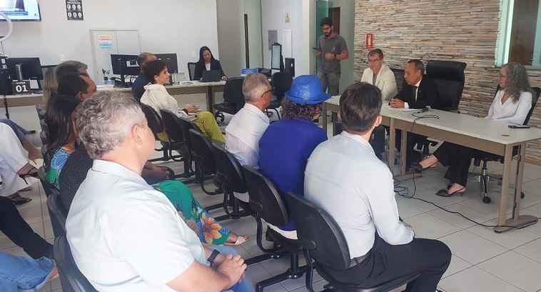 A imagem mostra uma reunião formal em uma sala equipada com mesas, cadeiras e computadores. Um g...
