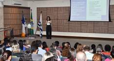 A imagem mostra um auditório com várias pessoas sentadas, acompanhando uma apresentação. No palc...