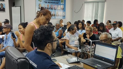 A imagem mostra uma sala cheia de pessoas, em um atendimento coletivo. No primeiro plano, um ser...