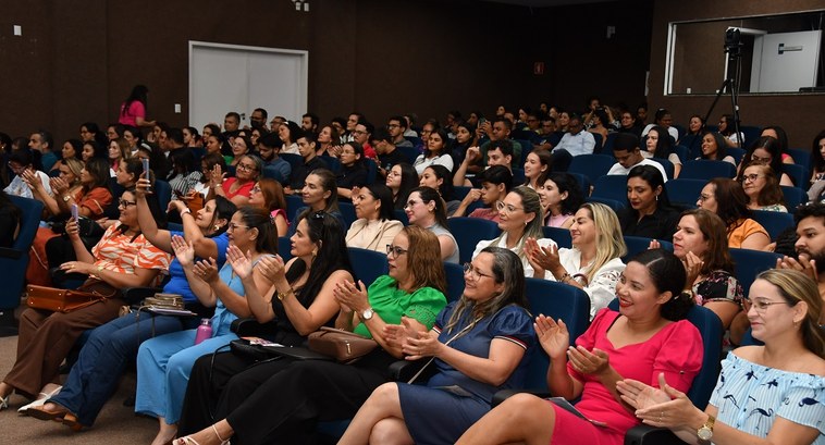 A imagem mostra um auditório cheio de pessoas sentadas, assistindo ao evento da Semana da Mulher...