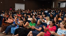A imagem mostra um auditório cheio de pessoas sentadas, assistindo ao evento da Semana da Mulher...