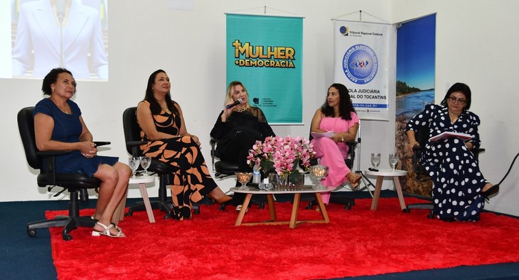 A imagem mostra um evento em ambiente fechado, com cinco mulheres sentadas em cadeiras sobre um ...