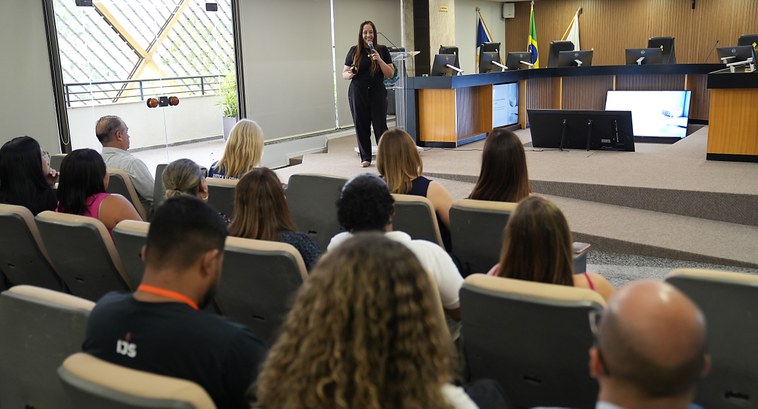 A imagem mostra uma mulher em pé, a palestrante Luciana Marquez, segurando um microfone e fazend...