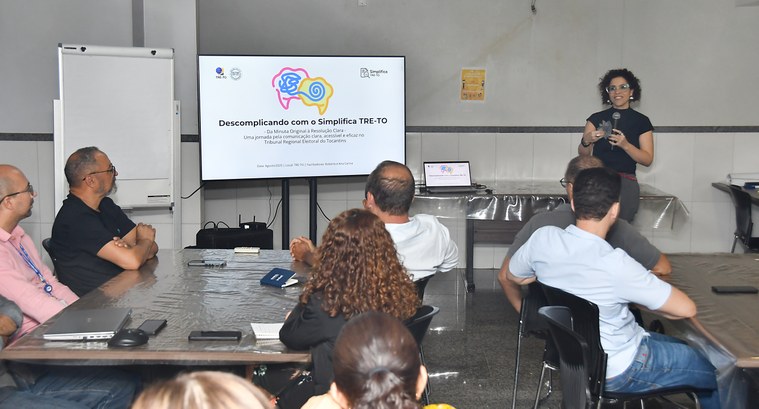 A imagem mostra uma sala onde acontece uma apresentação. À direita, a servidora Roberta Martins,...