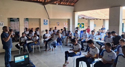 A imagem mostra uma palestra sendo ministrada em um ambiente escolar. Um homem em pé, à esquerda...