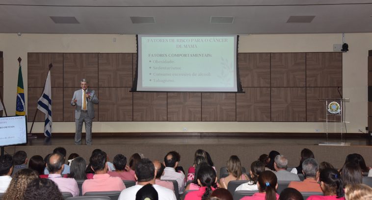 A imagem mostra a palestra realizada no auditório do TRE-TO. No palco, um homem de terno cinza e...