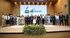 A imagem mostra um grupo de cerca de 25 pessoas posicionado sobre o palco de um auditório, duran...