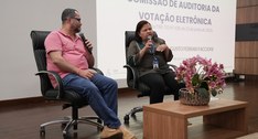 A imagem mostra dois participantes sentados em cadeiras giratórias sobre um palco de auditório d...