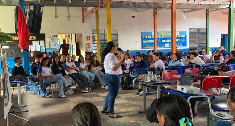 A imagem mostra uma palestra sendo realizada em um espaço escolar amplo e aberto, com vários est...