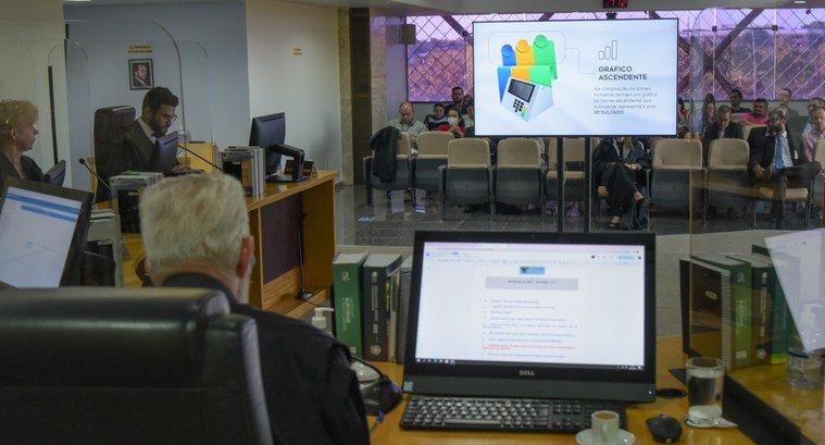 Foto da sessão plenária. Em uma TV o logo aparece com a informação "Gráfico ascendente. Na compo...