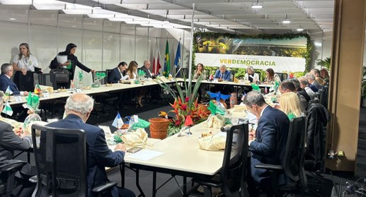 A imagem mostra uma reunião formal com várias pessoas sentadas ao redor de uma grande mesa em fo...