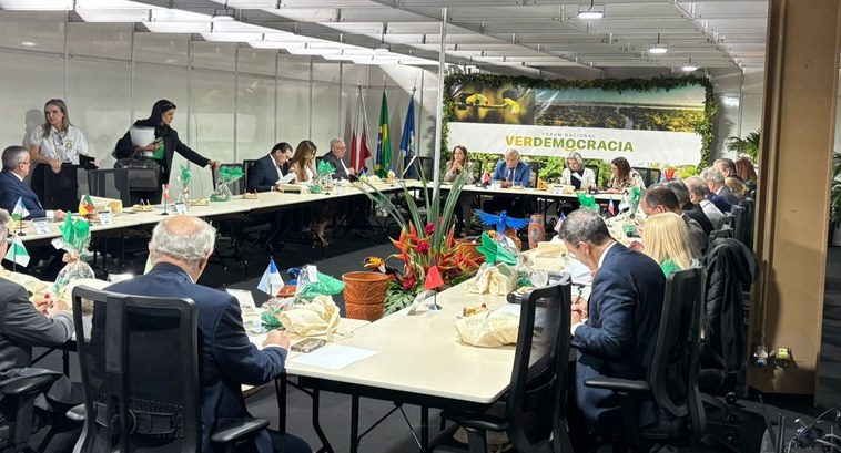 A imagem mostra uma reunião formal com várias pessoas sentadas ao redor de uma grande mesa em fo...