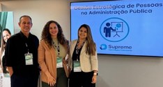 A imagem mostra três pessoas posando para uma foto em ambiente interno, em frente a uma tela que...
