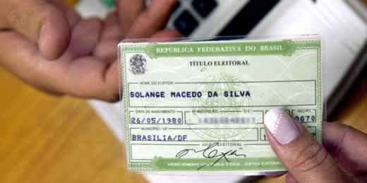 Título de Eleitor Mão feminina passando título eleitoral para mão masculina, com leitor biométrico ao fundo.