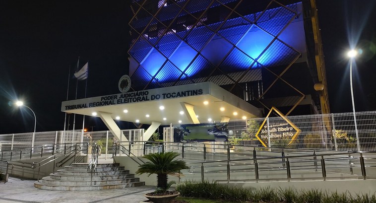 A imagem mostra a fachada do TRE-TO iluminada em tom azul durante a noite. O prédio, com sua est...