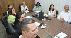 A imagem mostra um grupo de pessoas reunidas em torno de uma ampla mesa de madeira, em uma sala ...