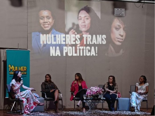 foto colorida da roda de conversa, com 5 mulheres sentadas lado a lado e com uma mesa com flores...