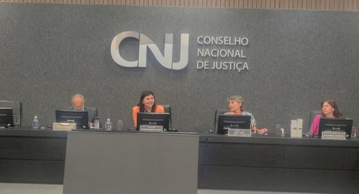 A imagem mostra uma reunião no CNJ, com quatro pessoas sentadas à mesa central, aparentemente di...