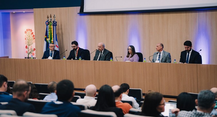 A imagem mostra um auditório durante um evento institucional. No palco, há uma mesa de madeira c...