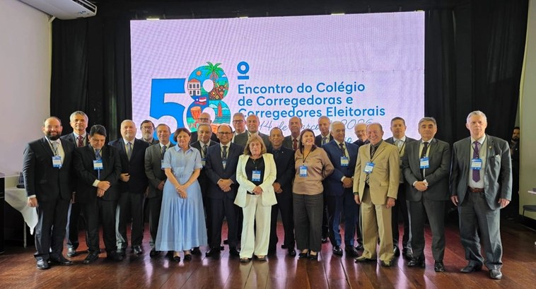 A imagem mostra um grupo de cerca de vinte pessoas reunidas em um ambiente interno, posando para...