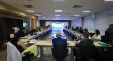 A imagem mostra uma reunião institucional realizada em uma sala ampla e moderna, com uma mesa em...