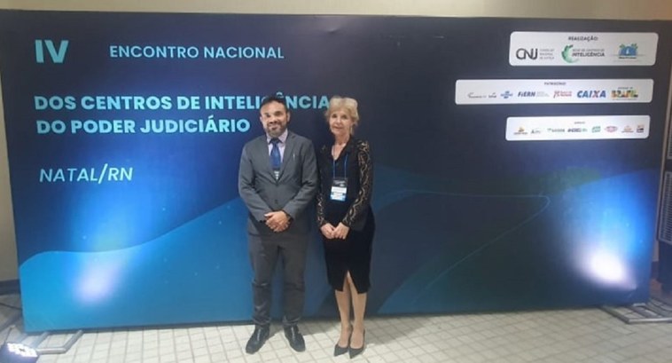 A imagem mostra duas pessoas posando para uma foto em frente a um painel institucional do IV Enc...