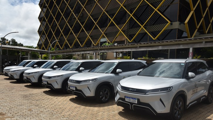 A imagem mostra uma fileira de veículos oficiais estacionados lado a lado no estacionamento inte...