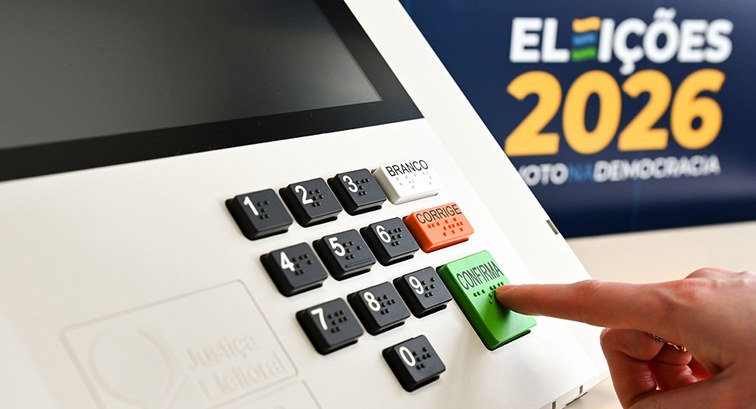 Foto em primeiro plano focada no teclado de uma urna eletrônica brasileira. Uma pessoa pressiona...