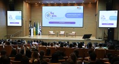 A imagem mostra um auditório lotado, com diversas pessoas sentadas assistindo a uma palestra. No...