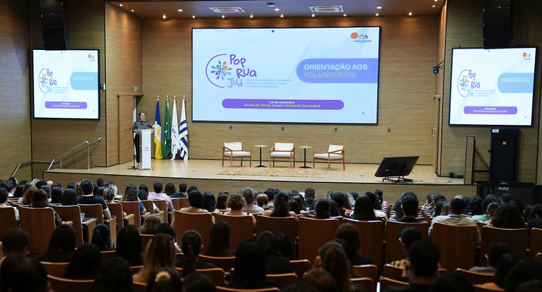 A imagem mostra um auditório lotado, com diversas pessoas sentadas assistindo a uma palestra. No...