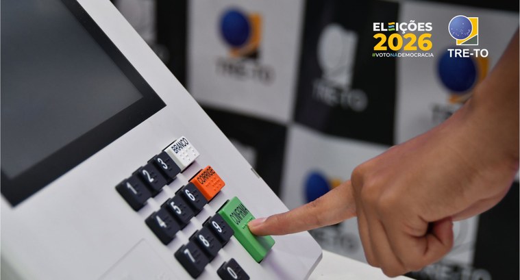A imagem mostra uma urna eletrônica em primeiro plano, com destaque para a mão de uma pessoa pre...