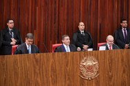 No plenário da sede do TSE, três homens estão sentados atrás de uma bancada de madeira que ocupa...
