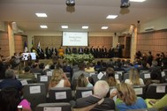 imagem colorida de evento de diplomação com autoridades no palco e diplomandos sentados no audit...